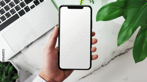 A hand holds up a blank smartphone screen, positioned over a marble desk adorned with a laptop and green plants.