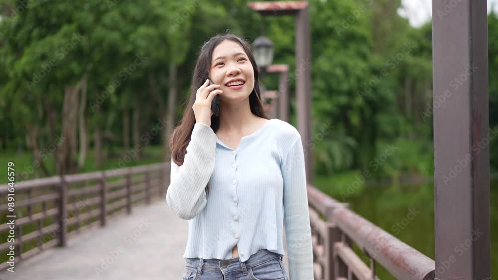 Asian woman standing on a bridge, smiling and talking on a phone, wearing a light blue top and jeans. The background is filled with lush green trees. Asian woman standing on a bridge, smiling and talking on a phone, wearing a light blue top and jeans. The background is filled with lush green trees.