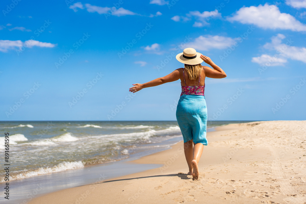 Summer vacation. Beautiful young woman in colorful swimsuit and blue pareo and hat walking on the sandy beach on a beautiful sunny day. Back view Summer vacation. Beautiful young woman in colorful swimsuit and blue pareo and hat walking on the sandy beach on a beautiful sunny day. Back view