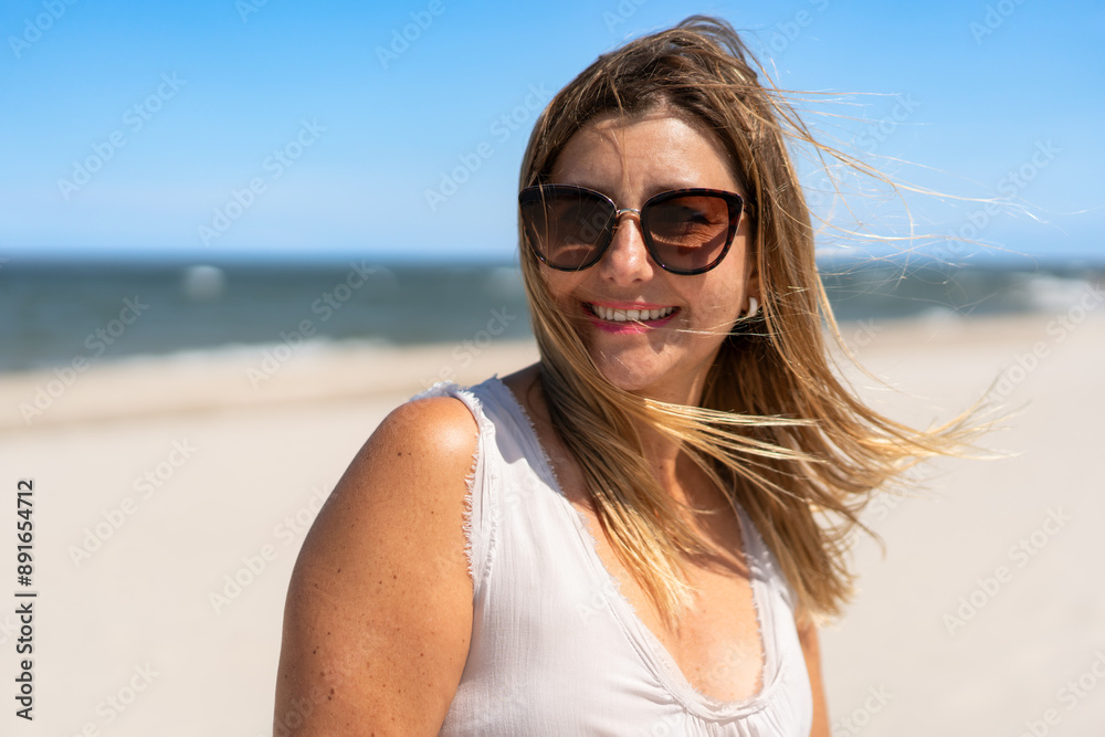 Summer. Portrait of young beautiful smiling woman with long blonde hair in light top and sunglasses against background of sandy beach and sea on sunny day. Energy, joy of life and youth