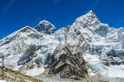 A view of Mount Everest, the world's tallest mountain, partially obscured by Lhotse, the fourth-tallest mountain.  Both peaks are snow-capped and tower over a rocky, brown landscape.