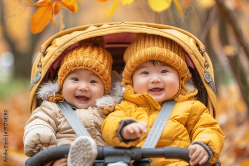 Charming Asian twin babies in yellow knitted hats and warm clothes are sitting in a stroller against the backdrop of an autumn park.