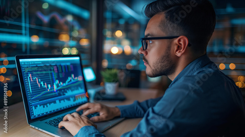 Young professional in glasses, sitting in his office, examining company charts and graphs on a laptop screen. Male employee analyzing corporate analytics at his workplace. © Maksym