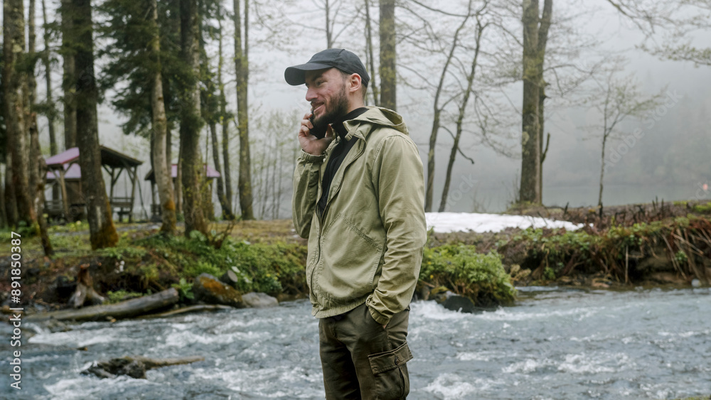 Obraz premium Tourist man talking on the phone near the river. Young hiker in forest camp, using mobile phone, traveling and hiking alone. 