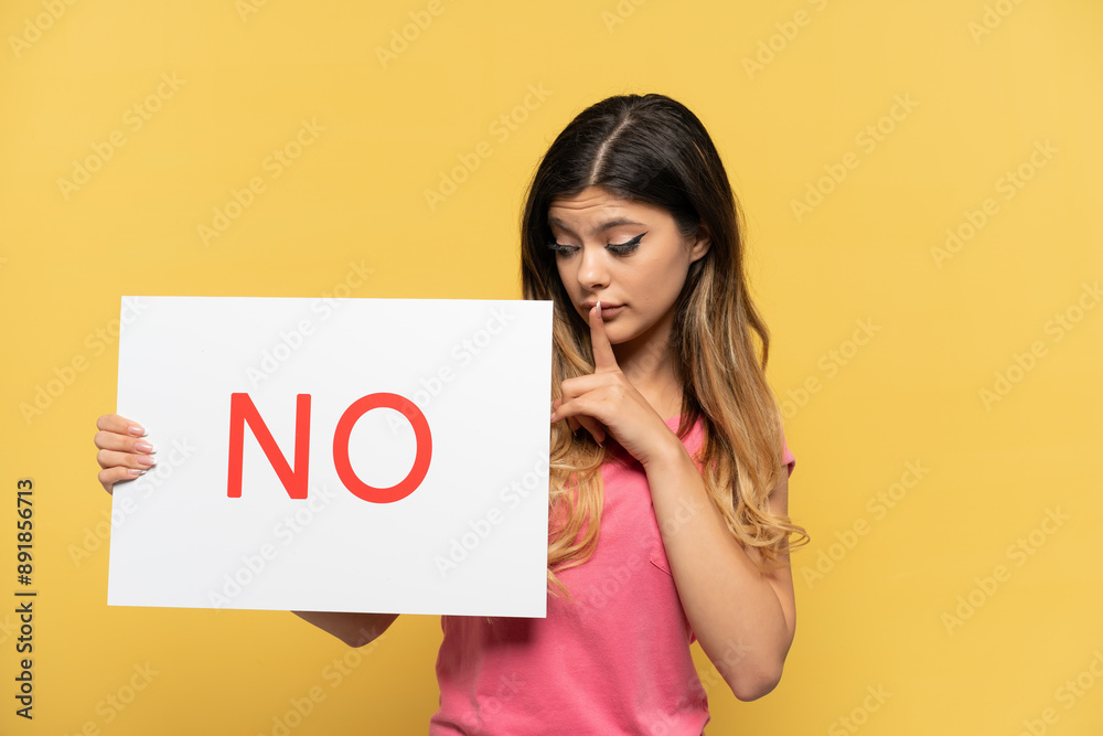 Young Russian girl isolated on yellow background holding a placard with text NO doing silence gesture Young Russian girl isolated on yellow background holding a placard with text NO doing silence gesture
