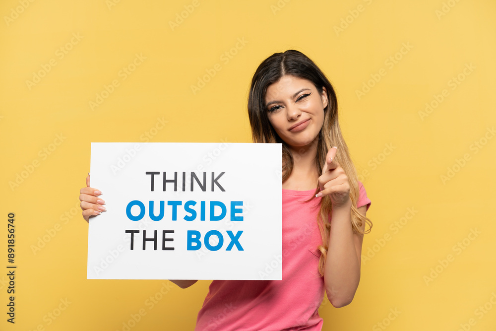 Young Russian girl isolated on yellow background holding a placard with text Think Outside The Box and pointing to the front Young Russian girl isolated on yellow background holding a placard with text Think Outside The Box and pointing to the front