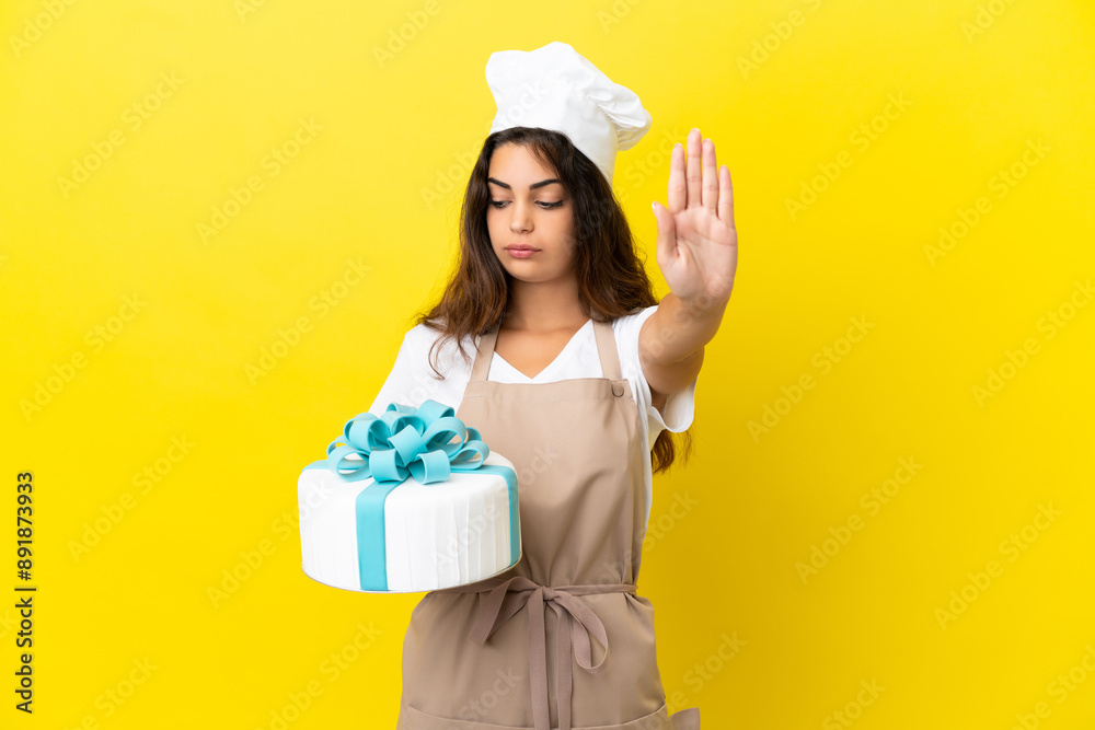 Young caucasian pastry chef woman with a big cake isolated on yellow background making stop gesture and disappointed Young caucasian pastry chef woman with a big cake isolated on yellow background making stop gesture and disappointed