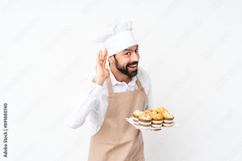 Young man holding muffin cake over isolated white background listening to something by putting hand on the ear Young man holding muffin cake over isolated white background listening to something by putting hand on the ear