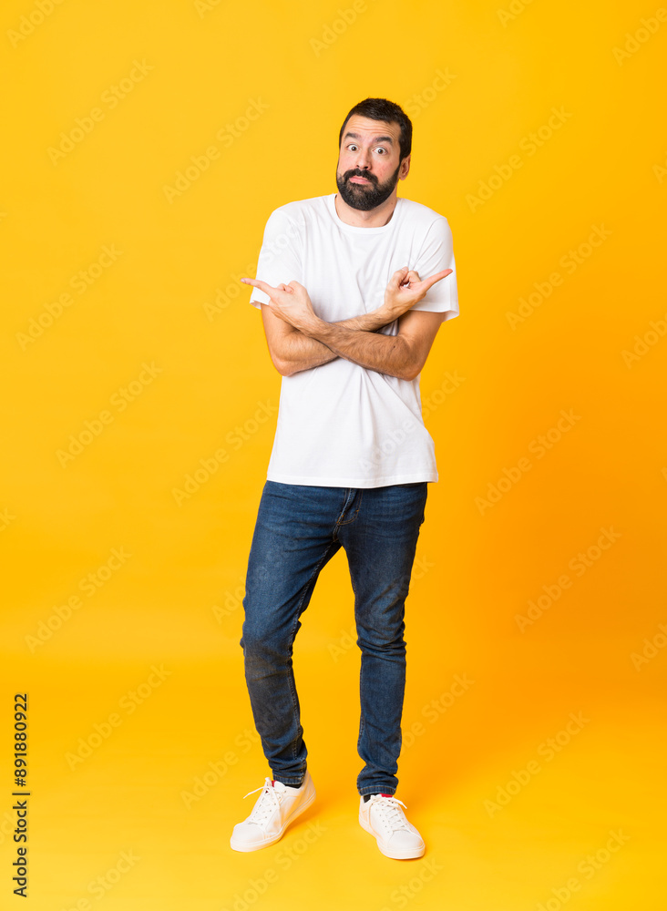 Full-length shot of man with beard over isolated yellow background pointing to the laterals having doubts Full-length shot of man with beard over isolated yellow background pointing to the laterals having doubts