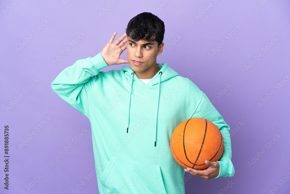 Young man playing basketball over isolated purple background listening to something by putting hand on the ear Young man playing basketball over isolated purple background listening to something by putting hand on the ear