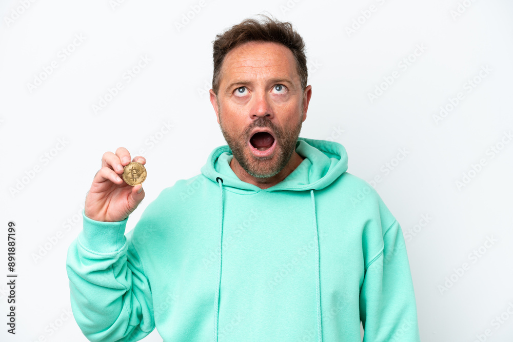 Middle age man holding a Bitcoin over isolated background looking up and with surprised expression