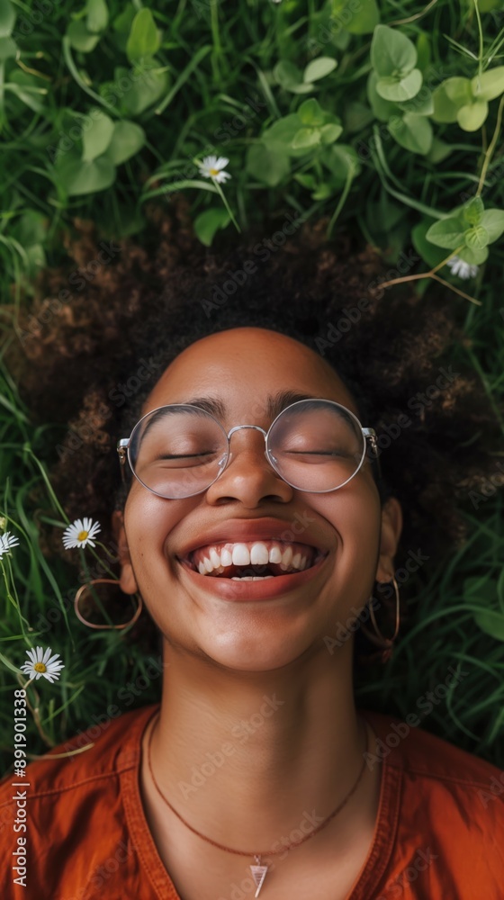 A Black woman with curly hair and glasses lies in grass, smiling up at the camera