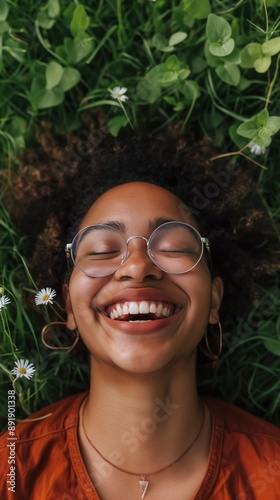 A Black woman with curly hair and glasses lies in grass, smiling up at the camera