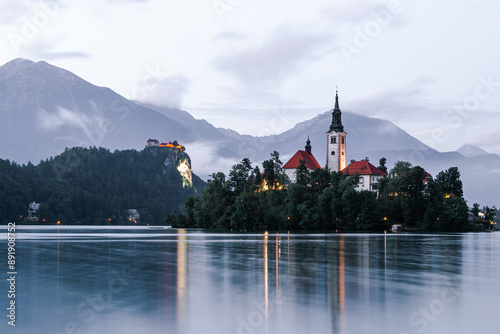 Bled, Slovenia (Slovenija), A chatolic church on the island in the lake by the mountainside with the castle on the big cliffed rock behind it in the evening hours with small partial clouds over them. photo