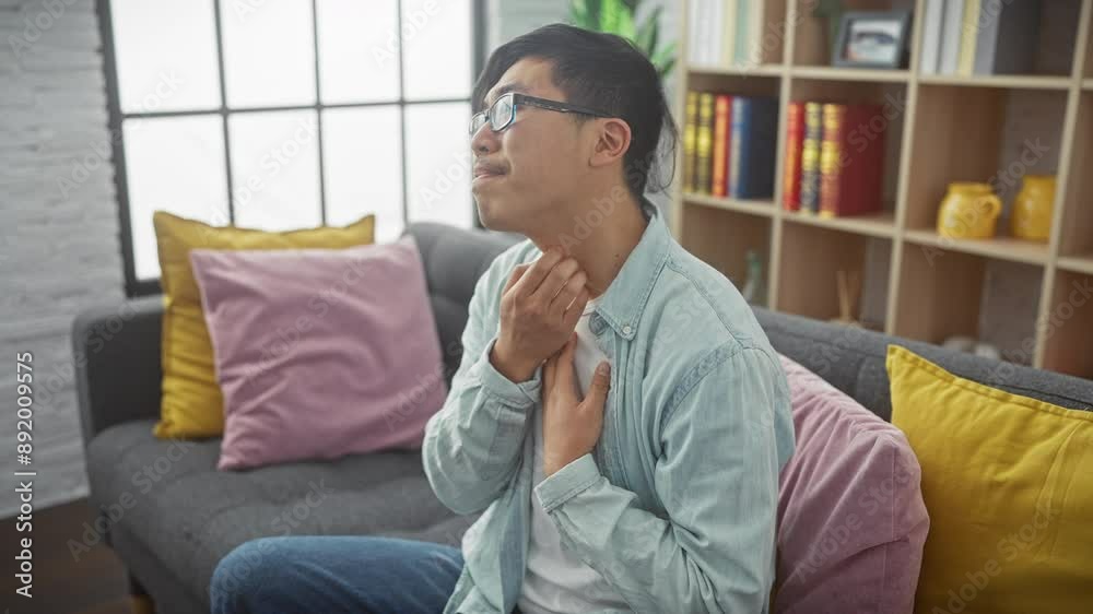 A young asian man scratches his neck while sitting on a couch in a modern living room, expressing discomfort or itching. A young asian man scratches his neck while sitting on a couch in a modern living room, expressing discomfort or itching.
