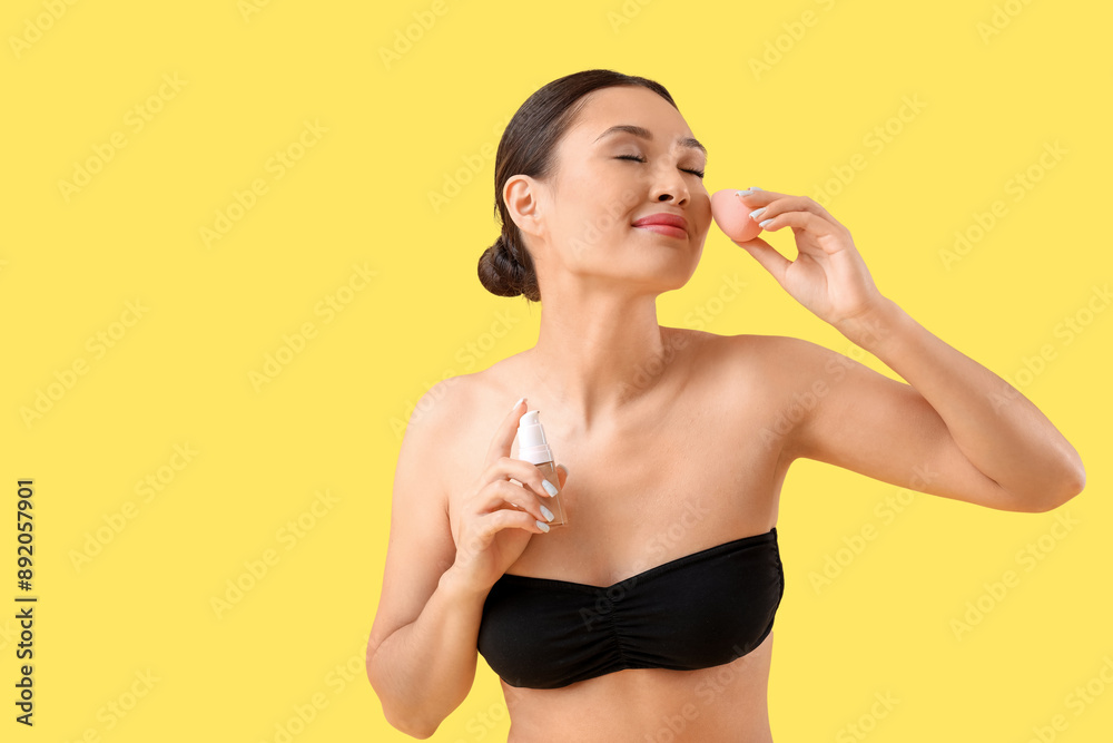 Young Asian woman applying makeup foundation with sponge on yellow background Young Asian woman applying makeup foundation with sponge on yellow background