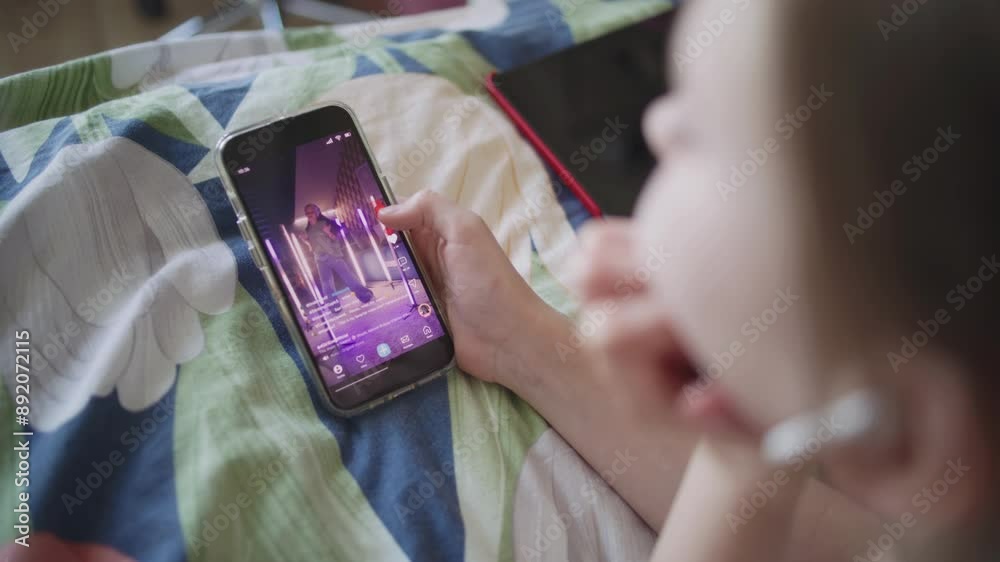 Caucasian girl surfs Internet spending leisure time in bedroom. Teenager lies on bed, watches video content in social network using mobile phone. Dancing young girl displayed on phone screen. Close up
