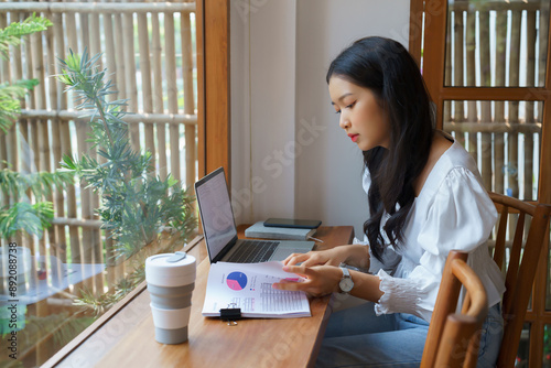 Young asian businesswoman checking marketing data of business on document and typing data on keyboard laptop while thinking about investment and planning of new startup project for working in cafe