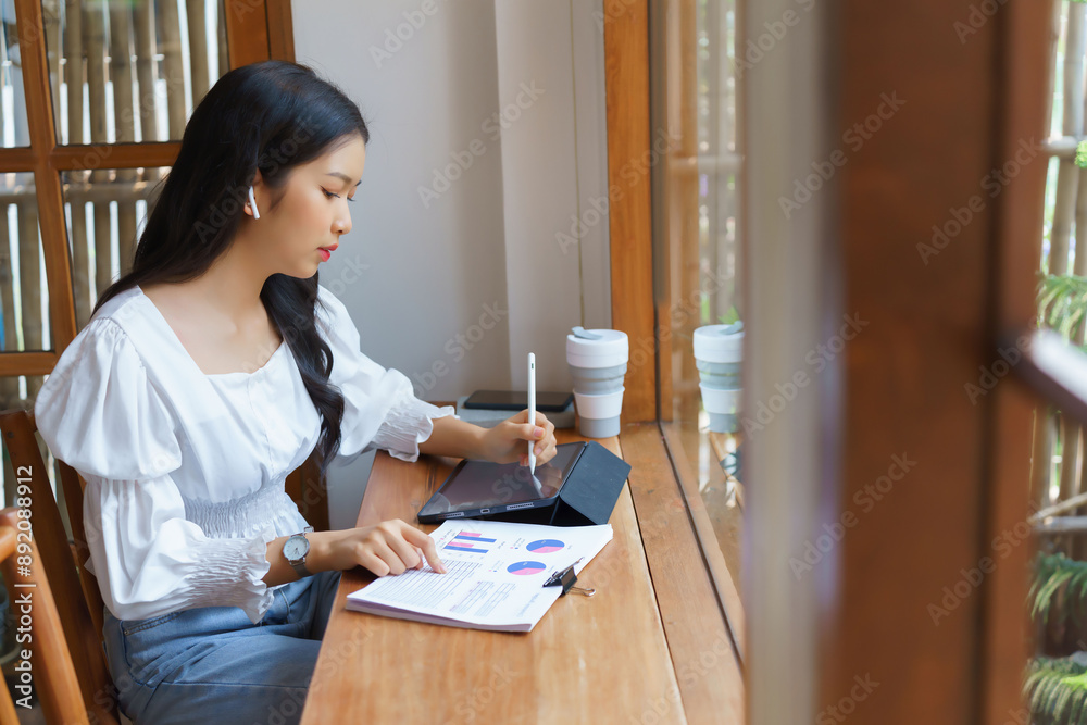 Young asian businesswoman reading business document and checking financial data to taking notes on tablet while thinking about investment and planning of new startup project to working in cafe Young asian businesswoman reading business document and checking financial data to taking notes on tablet while thinking about investment and planning of new startup project to working in cafe