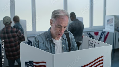Mature man comes to vote in booth in polling station office. National Election Day in the United States. Political races of US presidential candidates. Patriotism and civic duty concept. Dolly shot. photo
