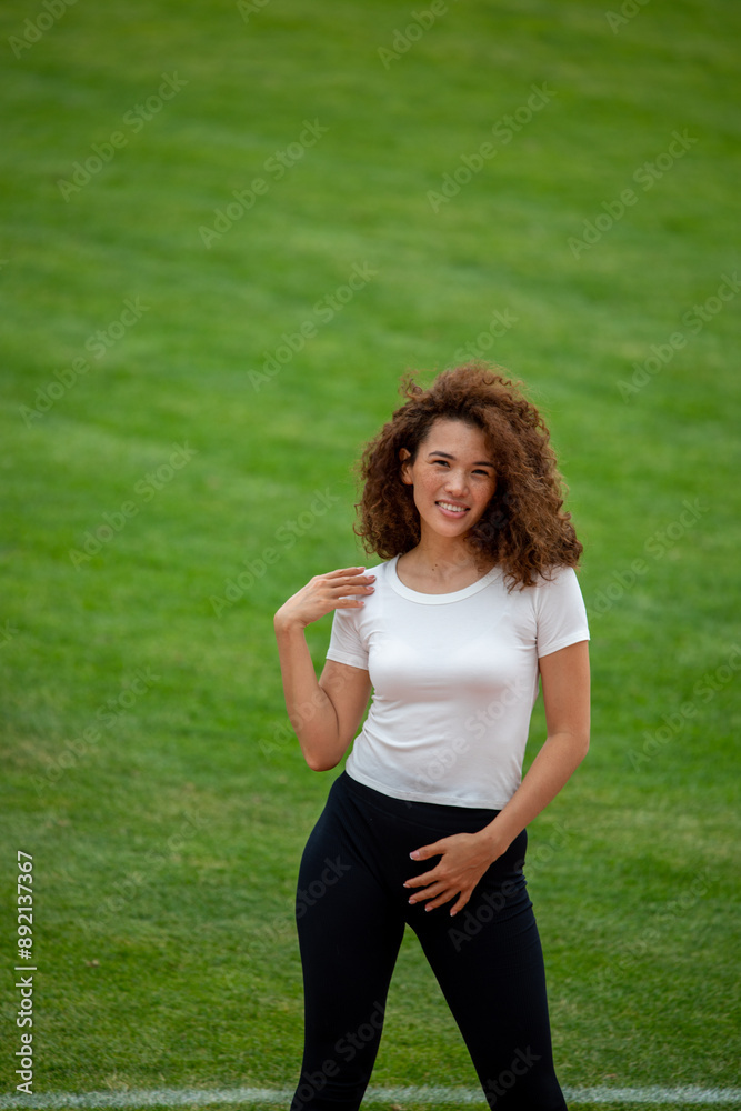 A beautiful curly girl posing in a white T-shirt and black leggings. Against the background of the stadium. Suitable for mockup A beautiful curly girl posing in a white T-shirt and black leggings. Against the background of the stadium. Suitable for mockup