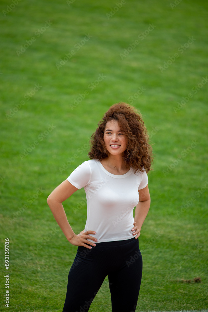 A beautiful curly girl posing in a white T-shirt and black leggings. Against the background of the stadium. Suitable for mockup A beautiful curly girl posing in a white T-shirt and black leggings. Against the background of the stadium. Suitable for mockup