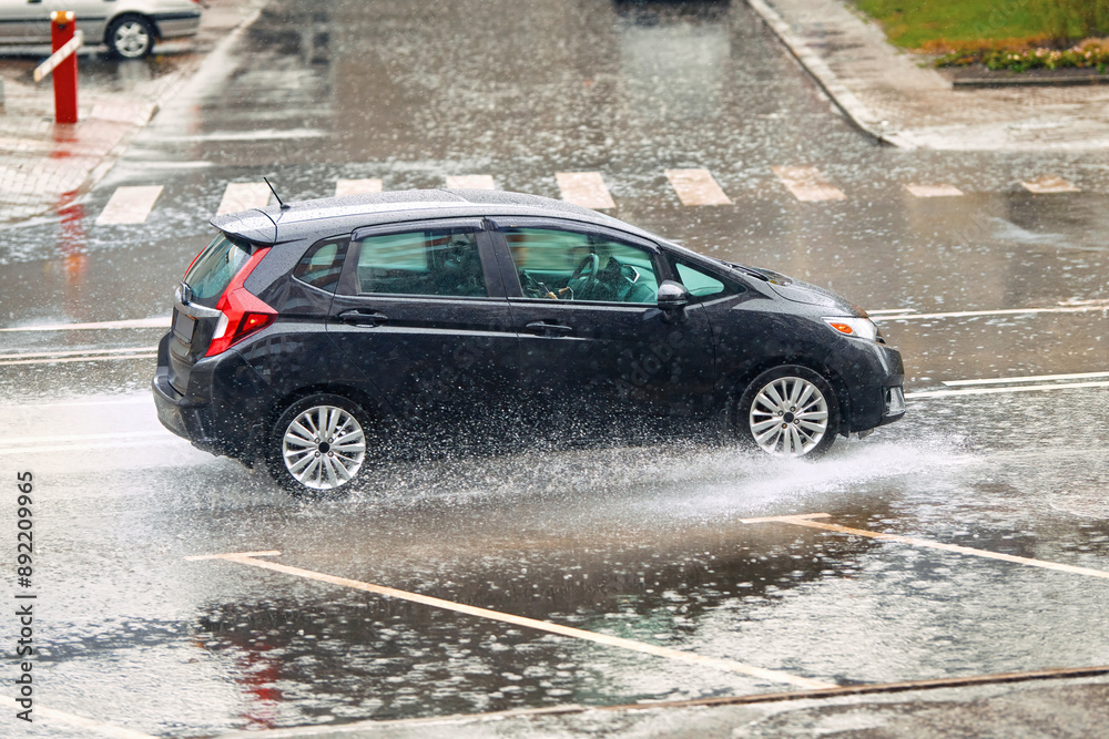 Compact car driving fast on wet road during rain, splashing water. Flood on the road, risk of hydroplaning. Car splashing through puddle on flooded road Compact car driving fast on wet road during rain, splashing water. Flood on the road, risk of hydroplaning. Car splashing through puddle on flooded road