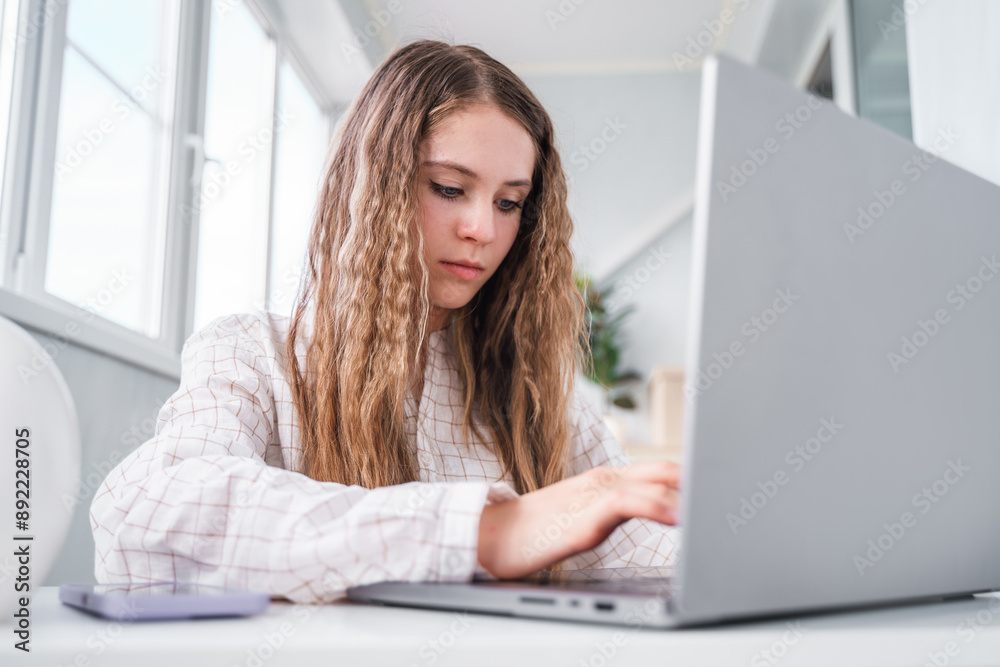 A young teenage student girl looking a laptop screen, online courses, studying remotely from home, sitting at a desk with a phone and laptop in a modern bright interior with plants. A young teenage student girl looking a laptop screen, online courses, studying remotely from home, sitting at a desk with a phone and laptop in a modern bright interior with plants.