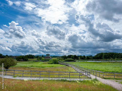 Grass and wetland area with wooden walk paths and bird viewing tower in distance. Dramatic clouds on summer day. No visible people photo