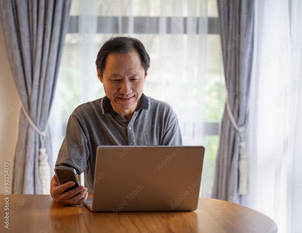 Man working on laptop, right hand holding a mobile phone at home office.