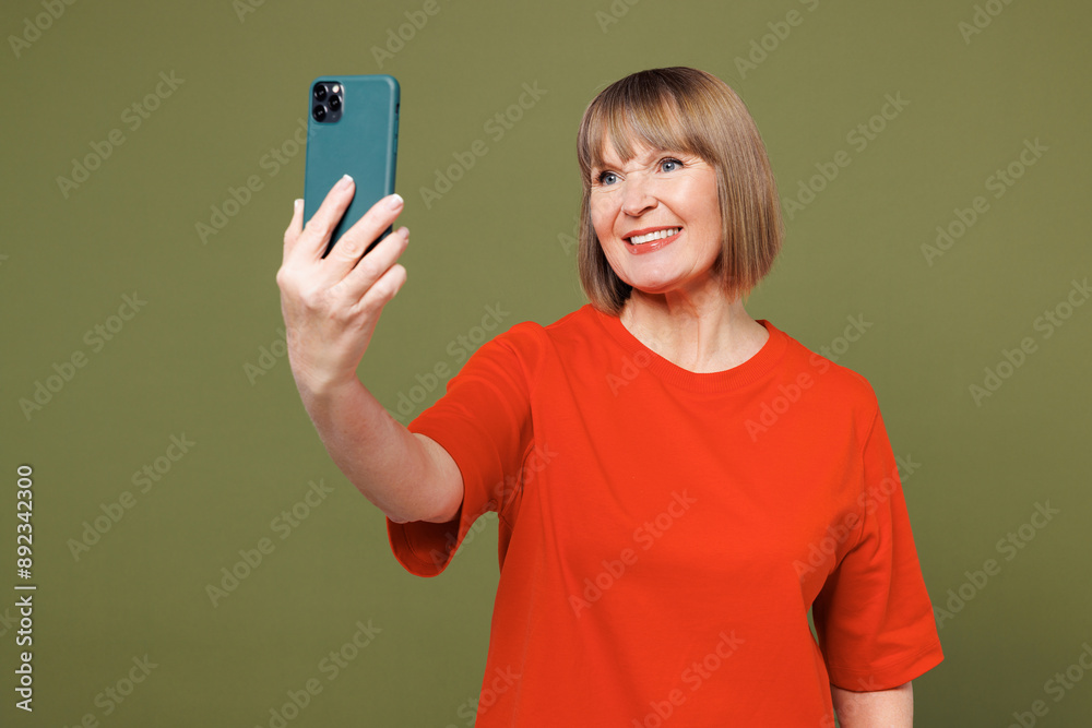 Elderly blonde woman 50s years old wear orange t-shirt casual clothes doing selfie shot on mobile cell phone post photo on social network isolated on plain pastel green background. Lifestyle concept. Elderly blonde woman 50s years old wear orange t-shirt casual clothes doing selfie shot on mobile cell phone post photo on social network isolated on plain pastel green background. Lifestyle concept.