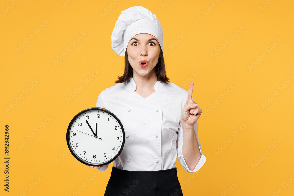 Young smart happy chef cook baker woman wear white shirt black apron uniform toque chefs hat hold in hand clock point index finger up isolated on plain yellow background studio. Cooking food concept. Young smart happy chef cook baker woman wear white shirt black apron uniform toque chefs hat hold in hand clock point index finger up isolated on plain yellow background studio. Cooking food concept.