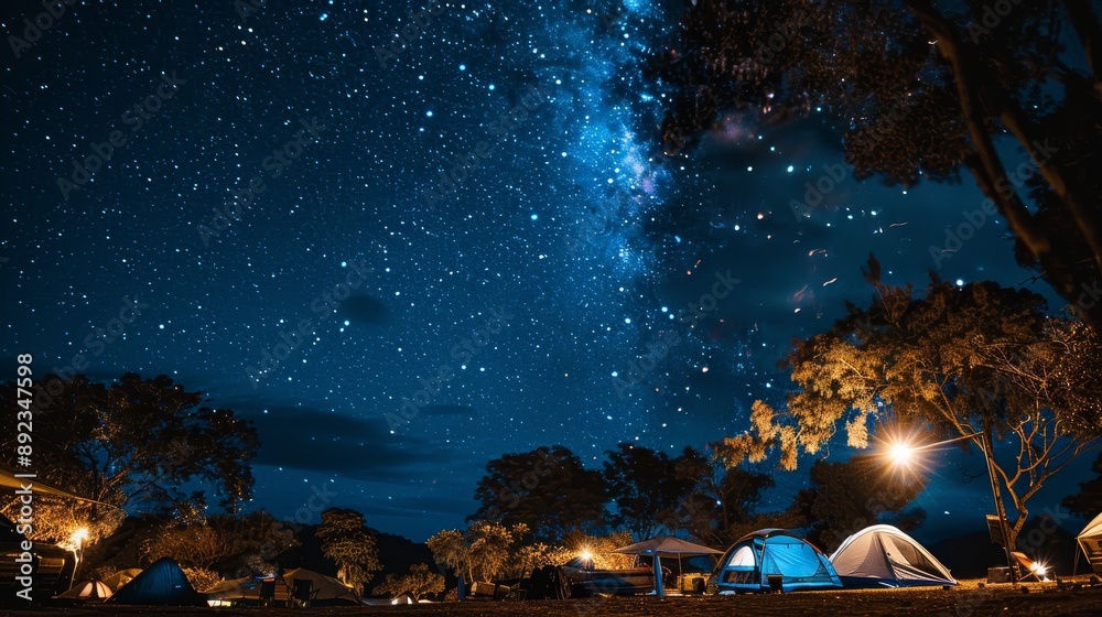 Obraz premium Starry night sky over a campsite with tents and trees illuminated by a warm light