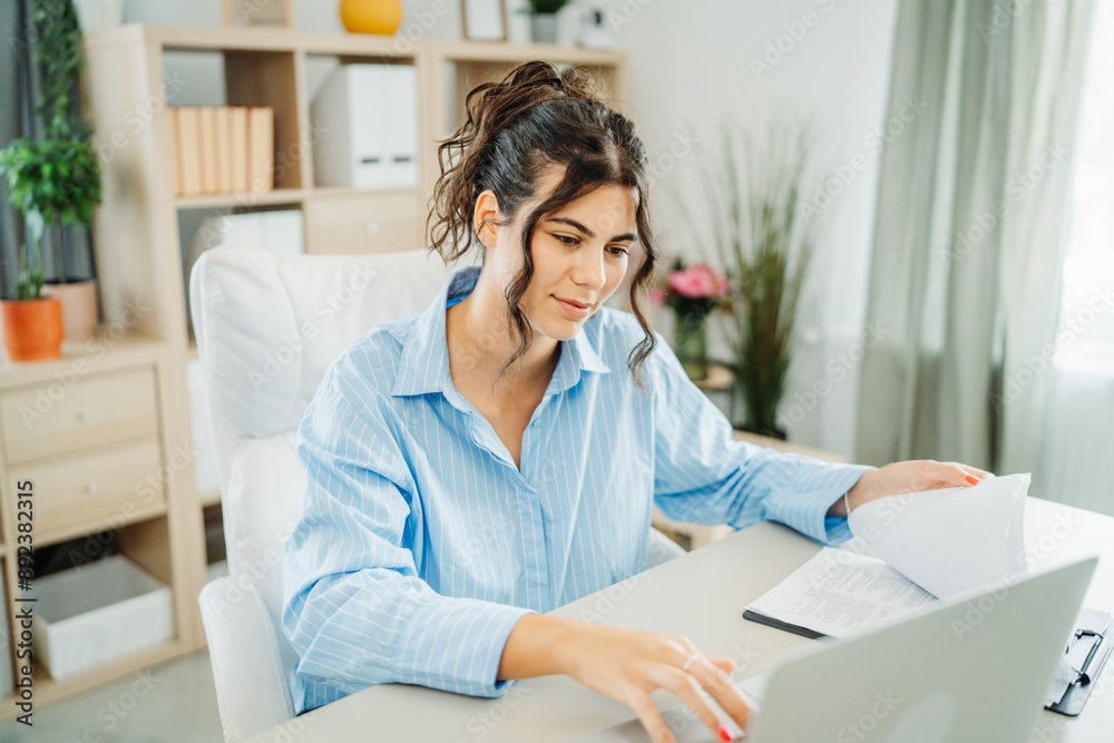 One young beautiful caucasian business woman working in office on laptop and looking through charts One young beautiful caucasian business woman working in office on laptop and looking through charts
