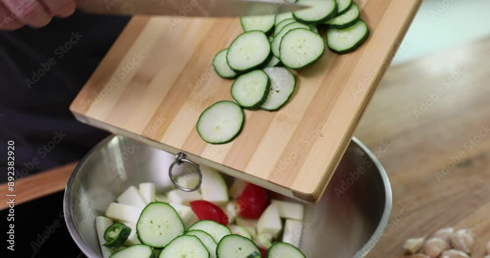 Man pushes sliced cucumber rings along wooden board with knife pouring into metal bowl. Alternately cutting salad ingredients slow motion