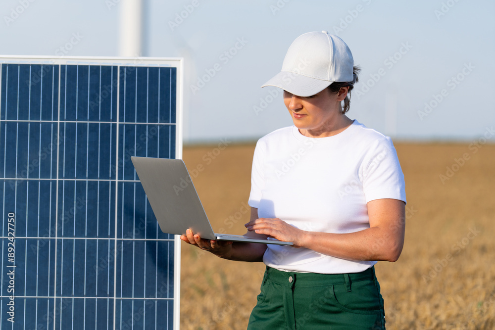 Woman farmer wearing white cap and t-shirt with laptop stands next to solar panel. Wind turbines in the background Woman farmer wearing white cap and t-shirt with laptop stands next to solar panel. Wind turbines in the background