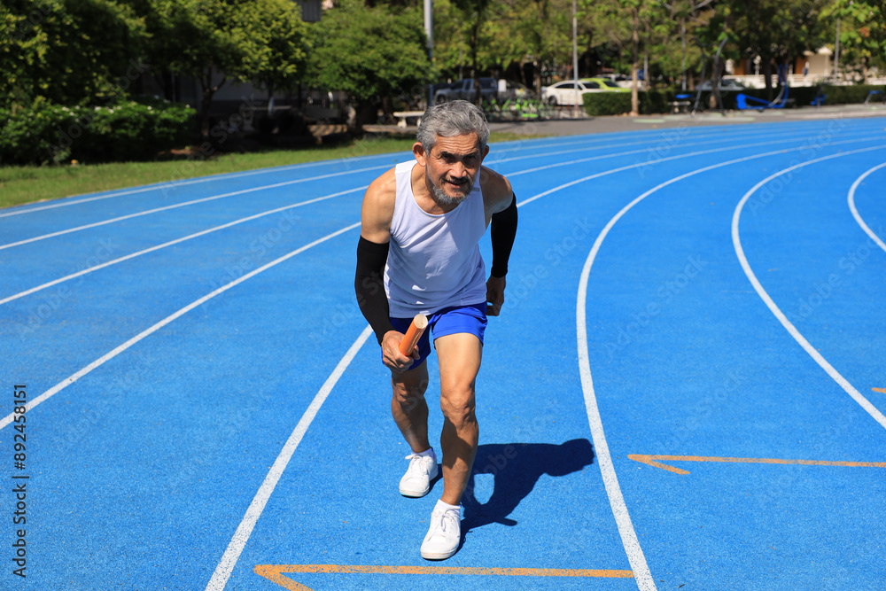 Obraz premium Senior Asian athlete is practicing running with relay baton in the professional blue track for professional exercise, sport training workout and healthy for longevity concept