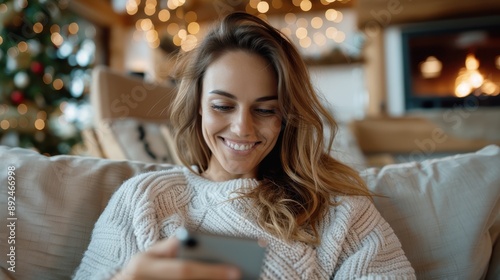 A woman smiling at her phone, sitting in a cozy space with festive decorations, wearing a warm sweater and enjoying a moment of relaxation and happiness during a holiday season.