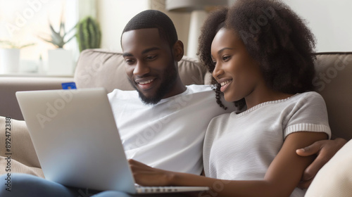 Husband and wife, a young African American couple, using a laptop and credit card for online shopping, sitting close on a comfortable sofa couch, enjoying their e-commerce experien © Maksym