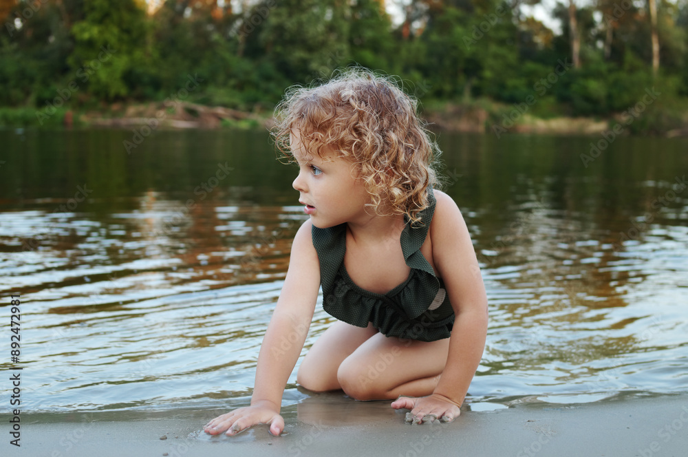 Blonde wavy haired Caucasian little girl wearing polka dot swimming suit sitting on river bank playing with sand while swimming in warm summer water Blonde wavy haired Caucasian little girl wearing polka dot swimming suit sitting on river bank playing with sand while swimming in warm summer water