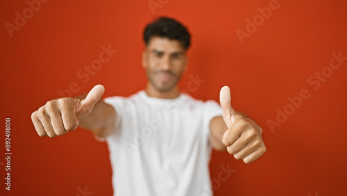 A young hispanic man gives a thumbs up against an isolated red background.