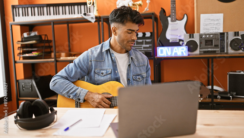 Handsome hispanic man playing guitar and using laptop in a modern music studio photo