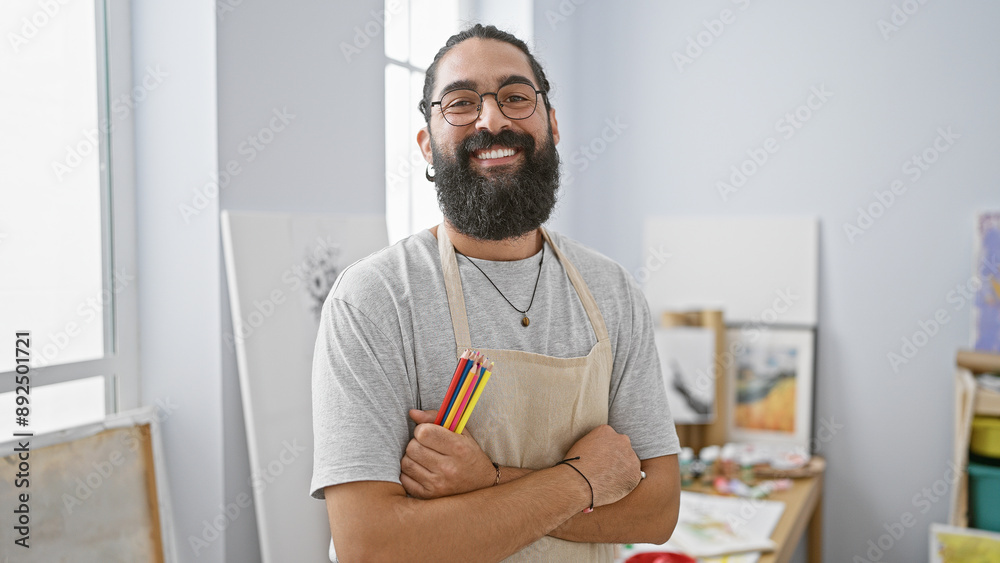 Fototapeta premium Hispanic artist with beard and apron smiling confidently indoors surrounded by paintings in a studio atmosphere.