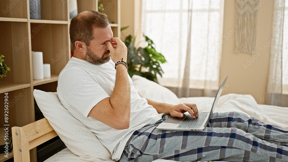 Mature man with grey hair experiences a headache while using a laptop in a cozy bedroom setting.