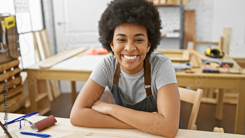 Portrait of a smiling black woman with curly hair, wearing a workshop apron indoors at her carpentry studio.