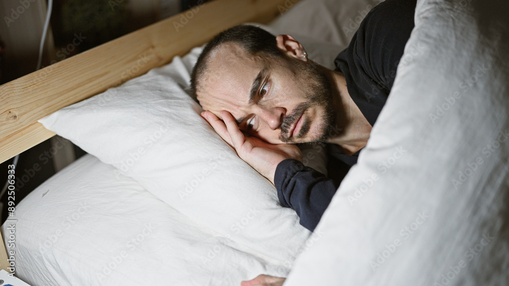 Hispanic man with beard lying in bed at home, looking tired or sleepless, in a dark bedroom setting at night. Hispanic man with beard lying in bed at home, looking tired or sleepless, in a dark bedroom setting at night.