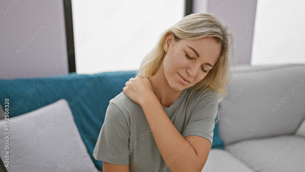 A beautiful blonde woman experiences discomfort in a modern living room, touching her neck gently. A beautiful blonde woman experiences discomfort in a modern living room, touching her neck gently.