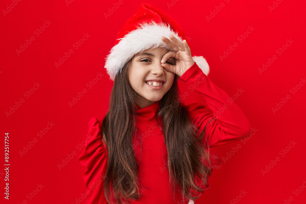 Adorable hispanic girl, confident and joyful, giving a fun 'ok' sign with fingers over eye. happy, smiling expression wearing christmas hat, isolated against a brilliant red background. Adorable hispanic girl, confident and joyful, giving a fun 'ok' sign with fingers over eye. happy, smiling expression wearing christmas hat, isolated against a brilliant red background.