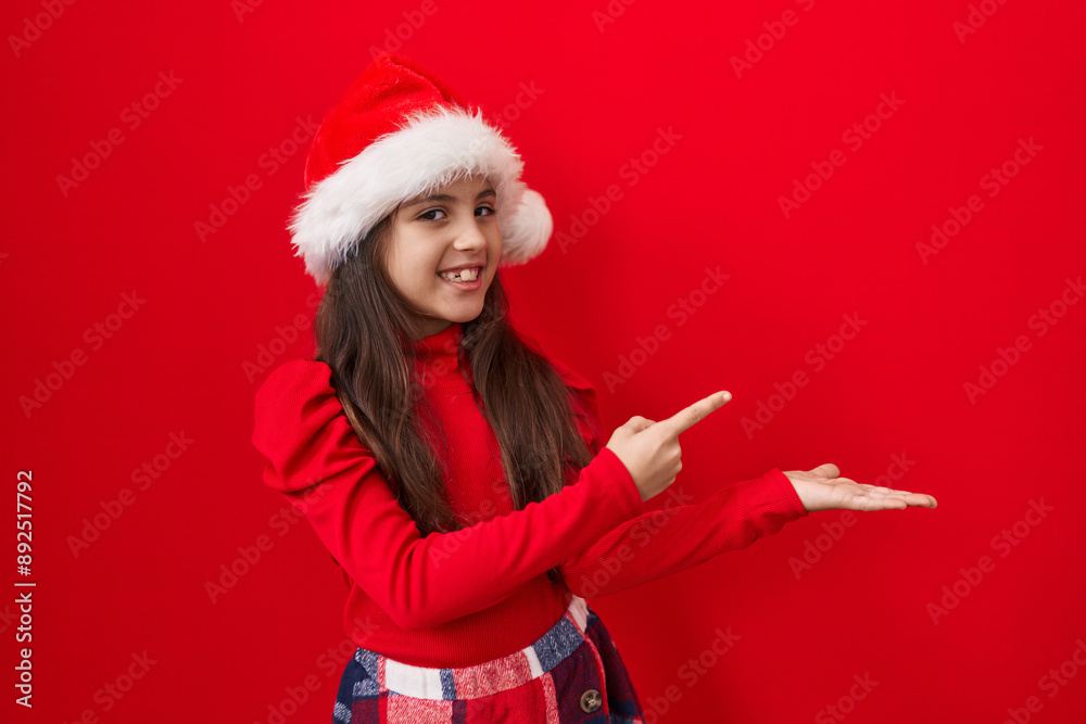 Adorable hispanic girl wearing christmas hat, cheerfully presenting and promoting product with confident hand gesture. smiling and pointing in amazement over isolated red background. Adorable hispanic girl wearing christmas hat, cheerfully presenting and promoting product with confident hand gesture. smiling and pointing in amazement over isolated red background.