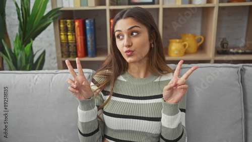 Fotografia A young hispanic woman poses playfully on a sofa indoors, flashing a peace sign with a coy expression